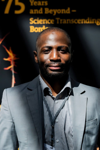 Adam Abdullahi stands in beams of sunlight for a headshot photo. Standing in front of a orange and black promotional sign.
