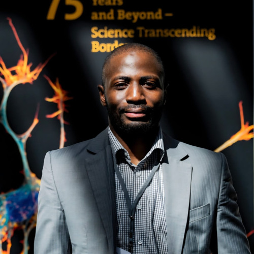 Adam Abdullahi stands in beams of sunlight for a headshot photo. Standing in front of a orange and black promotional sign.