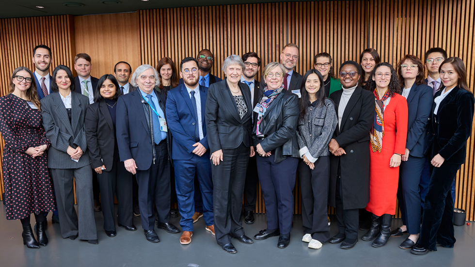 A group of 20 people stand closely together, smiling for the camera. They are in formal attire, set against a backdrop of vertical wooden panels, conveying a professional yet friendly atmosphere.