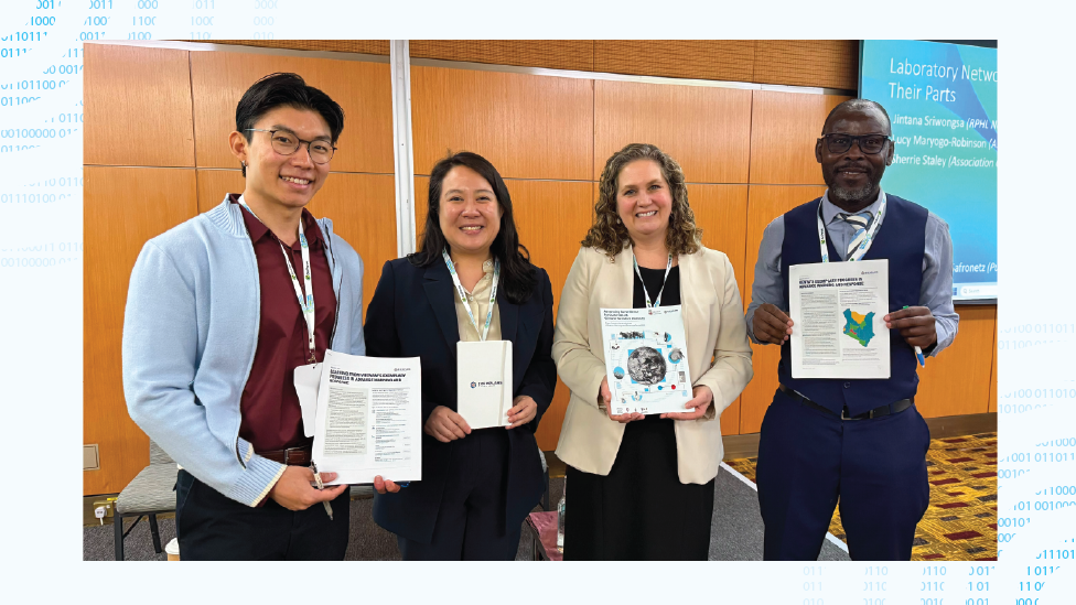 Phong Hong Khai, Anne Liu, Jennifer Nuzzo and Philip Ngere, stand on stage smiling and holding documents with a conference presentation in the background. 